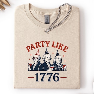 K&ouml;nnte beinhalten: Beiges T-Shirt mit dem Text "PARTY LIKE" &uuml;ber einer Illustration von drei historischen Figuren mit Partyh&uuml;ten und Bechern. Die Zahl "1776" ist darunter aufgedruckt. Eine silberne Halskette liegt &uuml;ber dem Shirt.