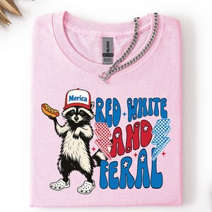 May include: A light pink t-shirt featuring a cartoon raccoon wearing a hat that says "Merica" and holding a hot dog. The shirt has the text "Red White and Feral" in red and blue. A silver chain necklace is draped across the shirt.