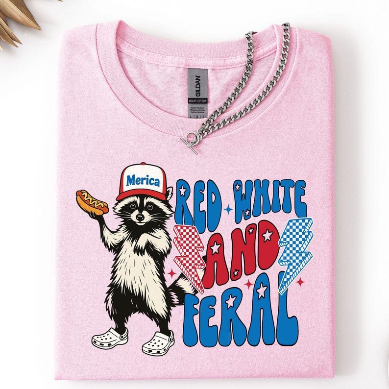 May include: A light pink t-shirt featuring a cartoon raccoon wearing a hat that says "Merica" and holding a hot dog. The shirt has the text "Red White and Feral" in red and blue. A silver chain necklace is draped across the shirt.