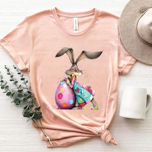 May include: A peach-colored t-shirt featuring a whimsical Easter design. The graphic depicts a gray bunny hugging a large, decorated egg. The egg is pink with colorful polka dots. The bunny wears a patchwork outfit.