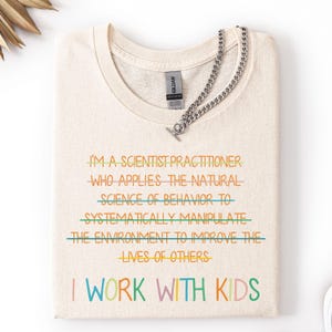 以下が含まれることがあります： さまざまな色で「I'M A SCIENTIST PRACTITIONER...I WORK WITH KIDS」というテキストがプリントされたクリーム色のTシャツ。トグルクラスプ付きのシルバーチェーンネックレスがシャツの上にかけられています。 シャツは折りたたまれています。
