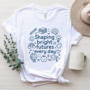 May include: White t-shirt with a navy blue graphic that reads "Shaping bright futures every day." The design includes a pencil, books, brain, lightbulb, sun, rainbow, globe, apple, graduation cap, and stars. A neutral-colored hat and white mug are in the background.