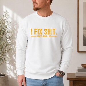 May include: A white long-sleeve crewneck sweatshirt featuring the yellow text "I FIX SHIT." with a wrench icon replacing the letter "I" in "SHIT," followed by the text "THAT'S WHAT I DO" underlined by a horizontal line.