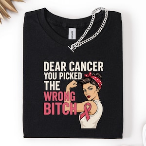 Puede incluir: Camiseta negra con el texto "DEAR CANCER YOU PICKED THE WRONG BITCH" y una ilustración de una mujer flexionando su brazo, con una diadema roja de lunares y una cinta rosa. Una cadena plateada está drapeada sobre la camiseta.