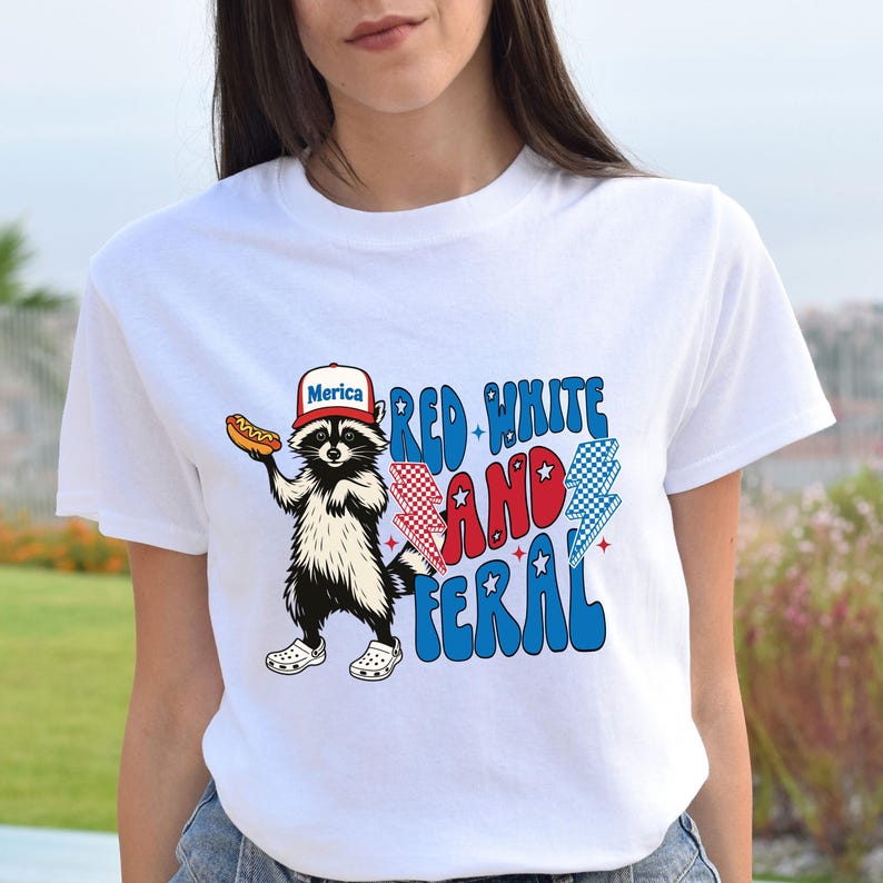 May include: White t-shirt featuring a raccoon wearing a "Merica" hat and holding a hot dog. The shirt has the text "Red, White, and Feral" in red, white, and blue colors with lightning bolt graphics. The raccoon is wearing white clogs.