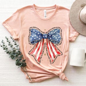 May include: A light pink t-shirt featuring a patriotic bow design. The bow has a blue section with white stars and red and white stripes. The text "God shed is grace on thee" is printed around the bow.