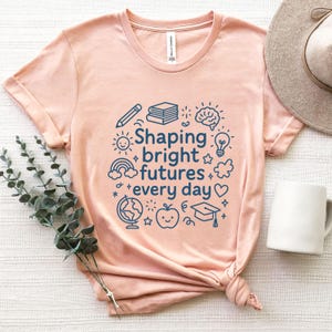 May include: A light pink t-shirt with the text "Shaping bright futures every day" in dark blue, surrounded by illustrations of a pencil, books, a brain, a lightbulb, and other school-related icons. The shirt is folded and knotted at the bottom.