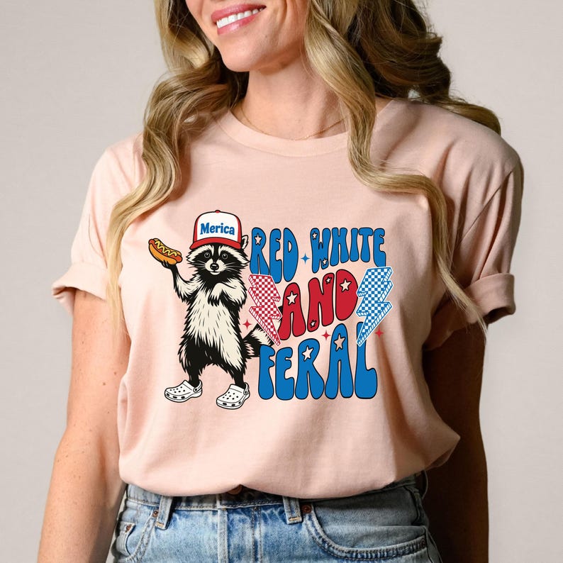 May include: A light pink t-shirt featuring a raccoon wearing a "Merica" hat and holding a hot dog. The shirt has the text "Red, White, and Feral" in red, white, and blue colors with lightning bolt graphics.