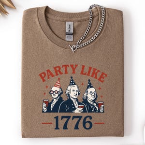 K&ouml;nnte beinhalten: Ein braunes T-Shirt mit Rundhalsausschnitt und der Aufschrift "PARTY LIKE" &uuml;ber einer Illustration von drei historischen Figuren, die Partyh&uuml;te tragen und Tassen halten. Die Zahl "1776" ist darunter gedruckt. Eine silberne Halskette liegt &uuml;ber dem Shirt.