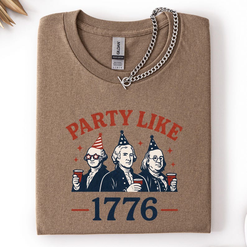K&ouml;nnte beinhalten: Ein braunes T-Shirt mit Rundhalsausschnitt und der Aufschrift "PARTY LIKE" &uuml;ber einer Illustration von drei historischen Figuren, die Partyh&uuml;te tragen und Tassen halten. Die Zahl "1776" ist darunter gedruckt. Eine silberne Halskette liegt &uuml;ber dem Shirt.