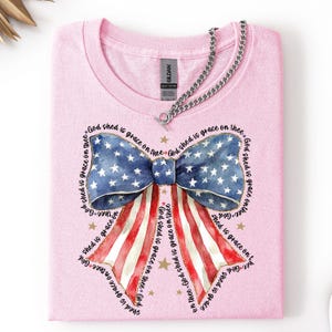 May include: A light pink t-shirt featuring a patriotic bow design. The bow incorporates the American flag with blue and white stars and red and white stripes. The text "God shed is grace on thee" surrounds the bow.