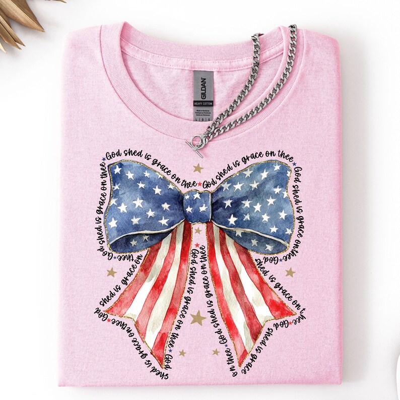May include: A light pink t-shirt featuring a patriotic bow design. The bow incorporates the American flag with blue and white stars and red and white stripes. The text "God shed is grace on thee" surrounds the bow.
