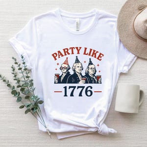 K&ouml;nnte beinhalten: Wei&szlig;es T-Shirt mit einer Grafik, die drei historische Pers&ouml;nlichkeiten mit Partyh&uuml;ten und roten Bechern zeigt. Der Text "PARTY LIKE" ist &uuml;ber den Figuren gew&ouml;lbt, darunter "1776" in Rot und Blau.