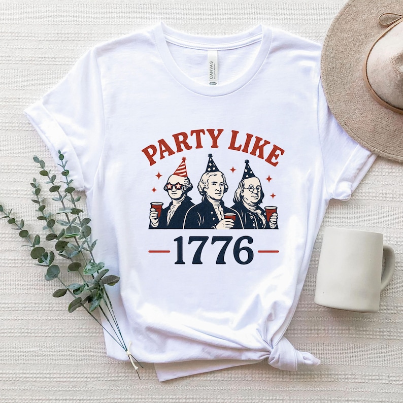 K&ouml;nnte beinhalten: Wei&szlig;es T-Shirt mit einer Grafik, die drei historische Pers&ouml;nlichkeiten mit Partyh&uuml;ten und roten Bechern zeigt. Der Text "PARTY LIKE" ist &uuml;ber den Figuren gew&ouml;lbt, darunter "1776" in Rot und Blau.