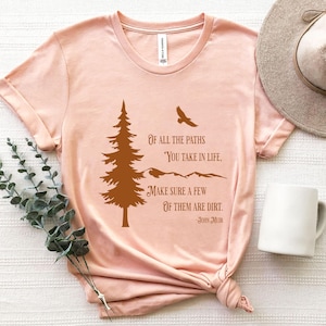 May include: A light pink t-shirt with a brown graphic of a pine tree, mountains, and a flying bird. The text reads "Of all the paths you take in life, make sure a few of them are dirt. -John Muir."