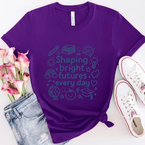 May include: A purple t-shirt with the text "Shaping bright futures every day" in teal. The design includes illustrations of a pencil, books, a brain, a lightbulb, a rainbow, a globe, an apple, and a graduation cap. White sneakers and blue jeans are also visible.