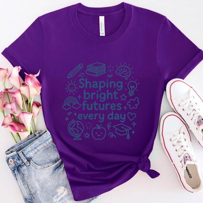 May include: A purple t-shirt with the text "Shaping bright futures every day" in teal. The design includes illustrations of a pencil, books, a brain, a lightbulb, a rainbow, a globe, an apple, and a graduation cap. White sneakers and blue jeans are also visible.