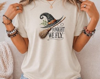Könnte beinhalten: Ein cremefarbenes Kurzarm-T-Shirt mit einer Grafik eines schwarzen Hexenhuts mit brauner Schnalle und einem Besen. Der Text "TONIGHT WE FLY" ist in schwarzen Buchstaben unter der Hut- und Besenillustration auf der Vorderseite aufgedruckt.