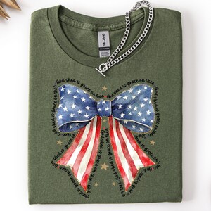 May include: An olive green t-shirt featuring a patriotic bow design. The bow incorporates the American flag's stars and stripes, with the text "God shed is grace on thee" encircling the bow. A silver chain necklace is draped across the shirt.