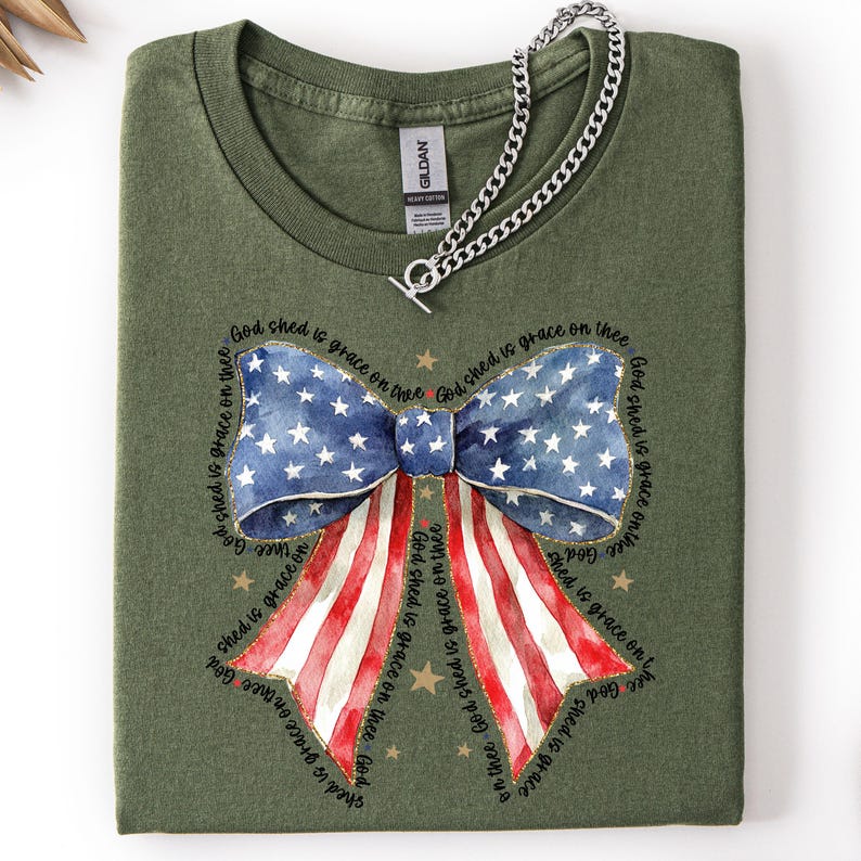 May include: An olive green t-shirt featuring a patriotic bow design. The bow incorporates the American flag's stars and stripes, with the text "God shed is grace on thee" encircling the bow. A silver chain necklace is draped across the shirt.