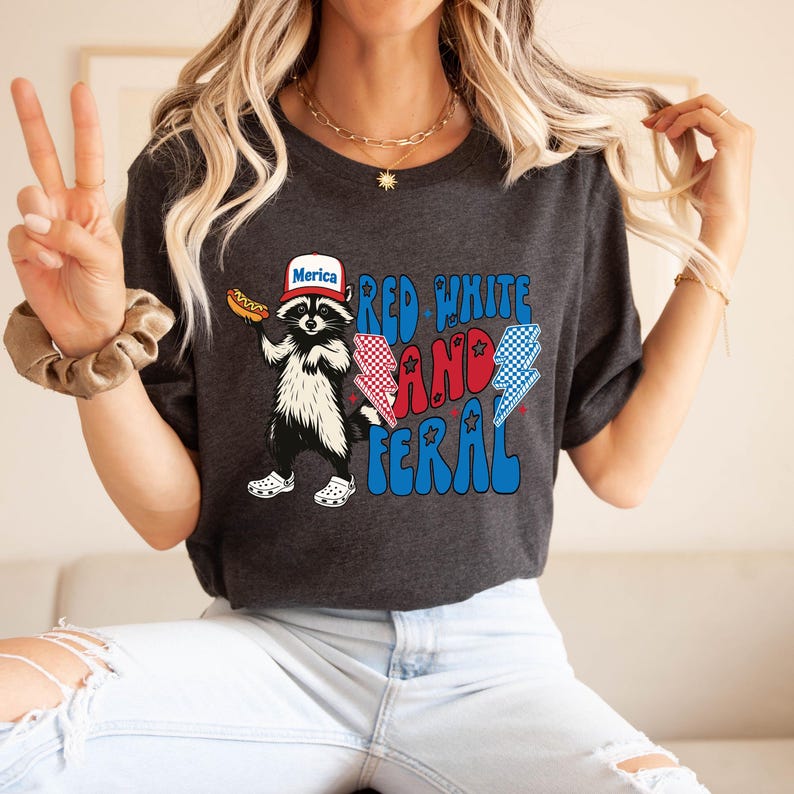 May include: Dark gray t-shirt featuring a raccoon wearing a hat that says "Merica" and holding a hot dog. The shirt has the words "Red White and Feral" with lightning bolt graphics. A fun and patriotic design.