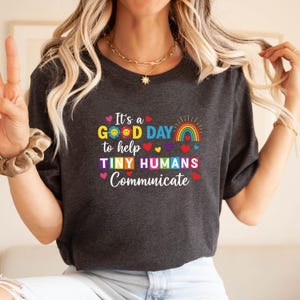 Puede incluir: Camiseta gris oscuro con el texto "It's a GOOD DAY to help TINY HUMANS Communicate" en letras coloridas. El diseño incluye un arcoíris, corazones y un sol sonriente. La camiseta tiene cuello redondo.