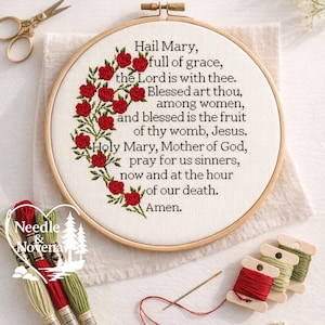 May include: A cross-stitch embroidery hoop with the text "Hail Mary" and a floral design of red roses. The embroidery is on a white fabric, with embroidery floss and a needle nearby. The Needle & Novena logo is in the lower left.