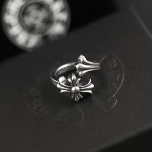 May include: A silver ring featuring an open-ended design with two ornate cross-shaped motifs. The metal has a polished finish with darkened recessed areas that highlight the intricate detailing of the cross emblems on each end of the band.