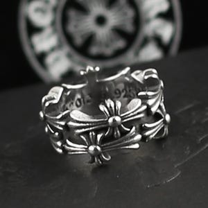 May include: A silver ring with a repeating cross design. The ring has a detailed, ornate pattern with a polished finish. The ring is likely made of sterling silver, as indicated by the "925" stamp.