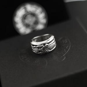 Puede incluir: Un anillo de plata con un diseño en relieve detallado de un motivo de daga o espada envuelto alrededor de la banda. El metal tiene un acabado pulido con áreas empotradas oscurecidas para resaltar los grabados intrincados de la superficie.