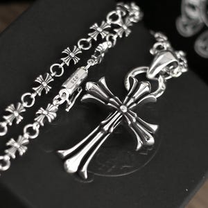 May include: A sterling silver necklace featuring a polished cross pendant with flared ends, suspended from a chain composed of repeating small cross-shaped links. A cylindrical clasp component is clearly engraved with the hallmark .925.