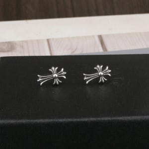 Op de afbeelding: Een paar zilverkleurige oorknopjes, elk voorzien van een gepolijst kruisontwerp met uitlopende, afgeronde uiteinden en een centraal cirkelvormig detail. De oorbellen hebben een metallic afwerking en zijn duidelijk zichtbaar.