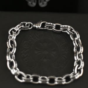 May include: A silver chain link bracelet featuring oval links with engraved cross motifs. The bracelet includes a lobster claw clasp with the engraving 925 and 2006. The metal has a polished finish with darkened recessed areas to highlight the details.