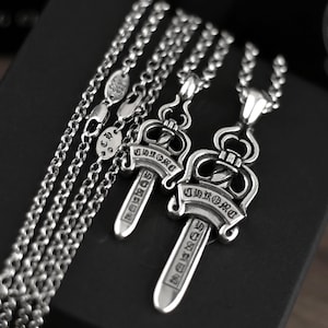 May include: Two silver-toned metal sword pendants hanging from matching link chains. Each pendant features an ornate, crown-like top and a blade engraved with the word CHROME. The chains include small oval tags stamped with 925 and 2001.