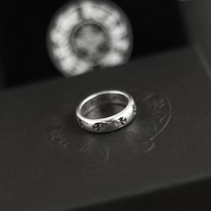 Op de afbeelding: Een gepolijste zilveren ring met een herhalend patroon van kleine, in reliëf aangebrachte kruismotieven rondom de buitenkant. De ring heeft een glad, afgerond profiel en een metallic afwerking die het licht reflecteert op het gedetailleerde ontwerp.