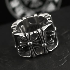 May include: A silver-toned ring with a square design. The ring features a repeating cross motif on the sides and top. The ring has a textured, detailed appearance, with a dark background.