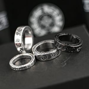 May include: Several silver rings with engraved lettering and decorative patterns. One ring is black with black stones. The rings are presented on a dark surface, highlighting their detailed designs and craftsmanship. The rings are a fashion accessory.