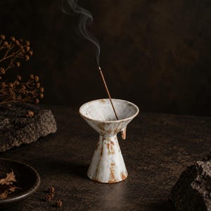 May include: A white ceramic incense holder with a flared, funnel-shaped top and a tapered base, featuring brown mottled accents. A single thin incense stick is placed inside the holder, emitting a visible trail of smoke rising upwards.