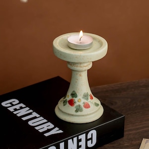 Puede incluir: Un portavelas de cerámica con una vela de té encendida. El portavelas es de color verde claro con una base de pedestal decorada con fresas rojas y hojas verdes. El portavelas está sobre un libro negro con la palabra "CENTURY" en blanco.