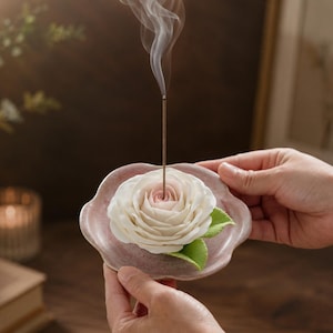 Puede incluir: Un porta-inciensos de cerámica con forma de plato festoneado en tono rosa empolvado, que presenta una rosa blanca esculpida con un centro rosa suave y hojas verdes. Una varilla de incienso está insertada en el centro de la rosa, emitiendo humo.
