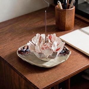May include: A ceramic incense holder shaped like a blooming flower with white and red-speckled petals, resting on a matching irregular tray with dark brown glazed edges. A thin, vertical incense stick is placed in the center, emitting smoke.
