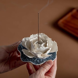 May include: A white ceramic incense holder shaped like a flower, with a dark blue base. The incense holder is in the shape of a flower with detailed petals. A lit incense stick is in the center, emitting a thin trail of smoke.