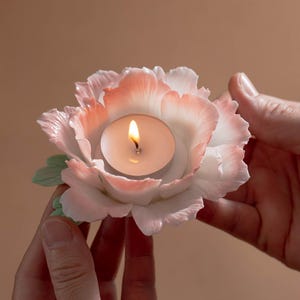 May include: A ceramic candle holder shaped like a peony flower, with white and pink petals. A lit tea light candle sits in the center. The flower has green leaves and is held by two hands.
