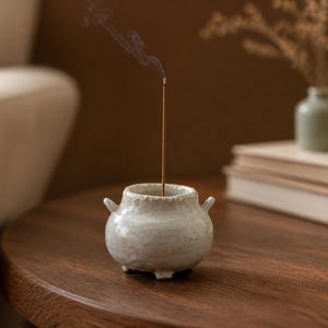May include: A small, round, off-white ceramic incense burner with a textured, speckled finish, two small side handles, and three short feet. A thin, brown incense stick is placed in the centre, emitting a thin trail of grey smoke upwards.