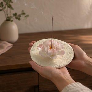 Puede incluir: Un porta-incienso circular de color blanco roto con textura y una flor de peonía rosa pálido esculpida en el centro. Un fino incienso de color marrón oscuro se mantiene erguido desde el centro de la flor, emitiendo un tenue hilo de humo.