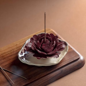 Può includere: Un porta incenso in ceramica bordeaux a forma di fiore, con un bastoncino di incenso acceso. Il supporto è su un vassoio di legno con bordo liscio e arrotondato. Il porta incenso ha un bordo bianco e viola.