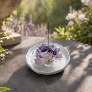 May include: A white ceramic incense holder featuring a sculpted purple and white flower at its center. A thin, dark incense stick is inserted into the middle of the flower, emitting a faint wisp of smoke that rises vertically into the air.