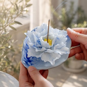 Puede incluir: Un porta-incienso de cerámica con forma de flor en floración, con pétalos superpuestos de color azul claro y blanco y un centro amarillo brillante. Un palillo de incienso marrón oscuro está insertado en el centro de la flor.
