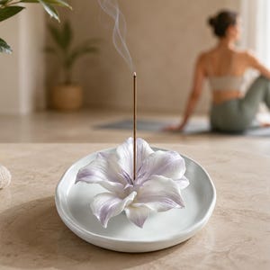 Puede incluir: Un porta-incienso de cerámica blanca con forma de flor en floración con suaves tonos morados en los pétalos. Una varilla de incienso marrón fina está colocada en el centro de la flor, emitiendo un fino rastro de humo que sube verticalmente.