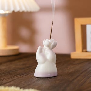 May include: A white ceramic incense holder, shaped like a figure with a lotus flower at the top. The base has a purple gradient. A lit incense stick is inserted, emitting a thin stream of smoke. The holder is on a wooden surface.